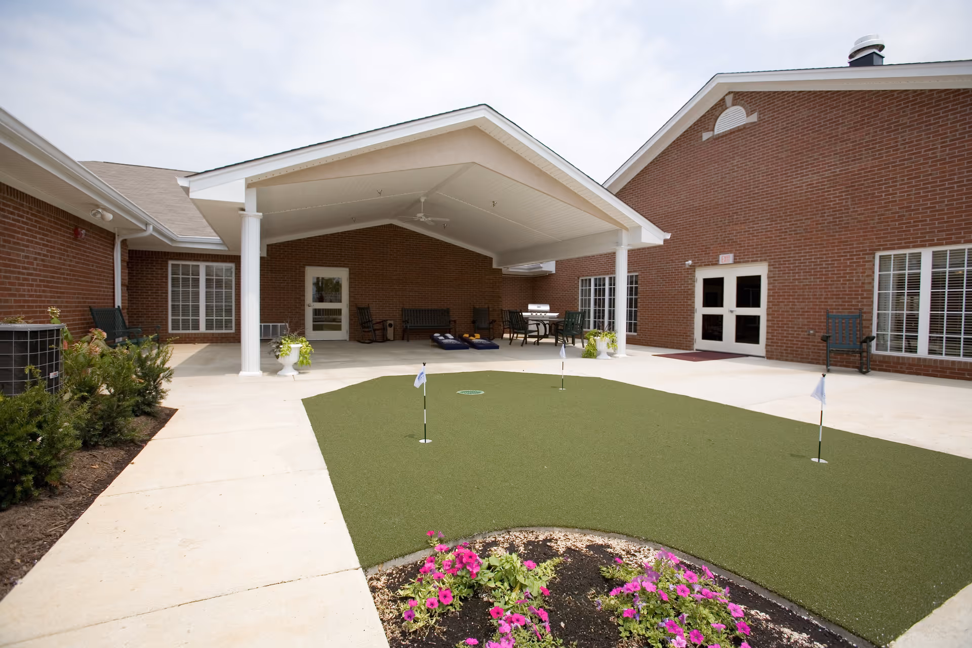 Outdoor courtyard area at Springhurst Health Campus featuring a small artificial putting green with three holes and flags, surrounded by a concrete walkway and flower beds with pink flowers. There is a covered patio with white columns, ceiling fans, rocking chairs, benches, a table with chairs, and a grill. The building has red brick walls and multiple windows and doors.