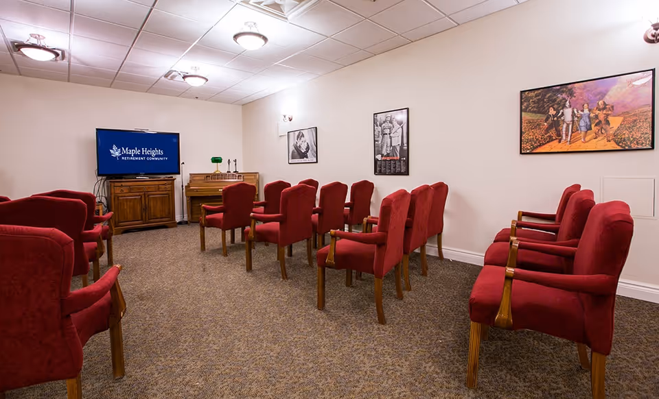 A small theater or presentation room with rows of red upholstered chairs facing a TV screen displaying the Maple Heights Retirement Community logo. The room has beige walls with framed pictures and a wooden cabinet beneath the TV. The ceiling has recessed lighting fixtures.