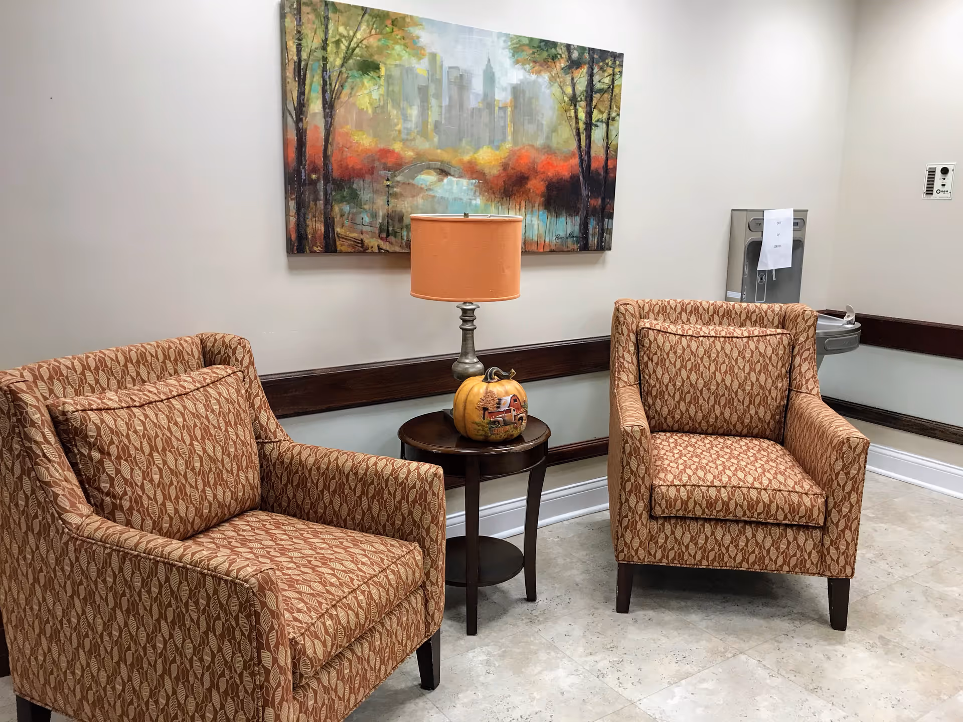 Two patterned armchairs with cushions are placed on either side of a small round wooden table with a lamp and a decorative pumpkin. A colorful painting of a park scene with trees and a city skyline hangs on the wall behind the chairs. A water fountain is visible on the right side of the image against a beige wall.