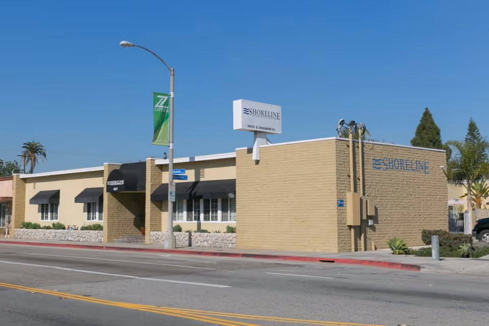 Exterior view of Shoreline Healthcare Center, a single-story beige brick building with black awnings over the windows and entrance. The building has signage displaying the name Shoreline and the address 4029 E. Anaheim St. A street lamp and a green banner are visible on the sidewalk in front of the building under a clear blue sky.