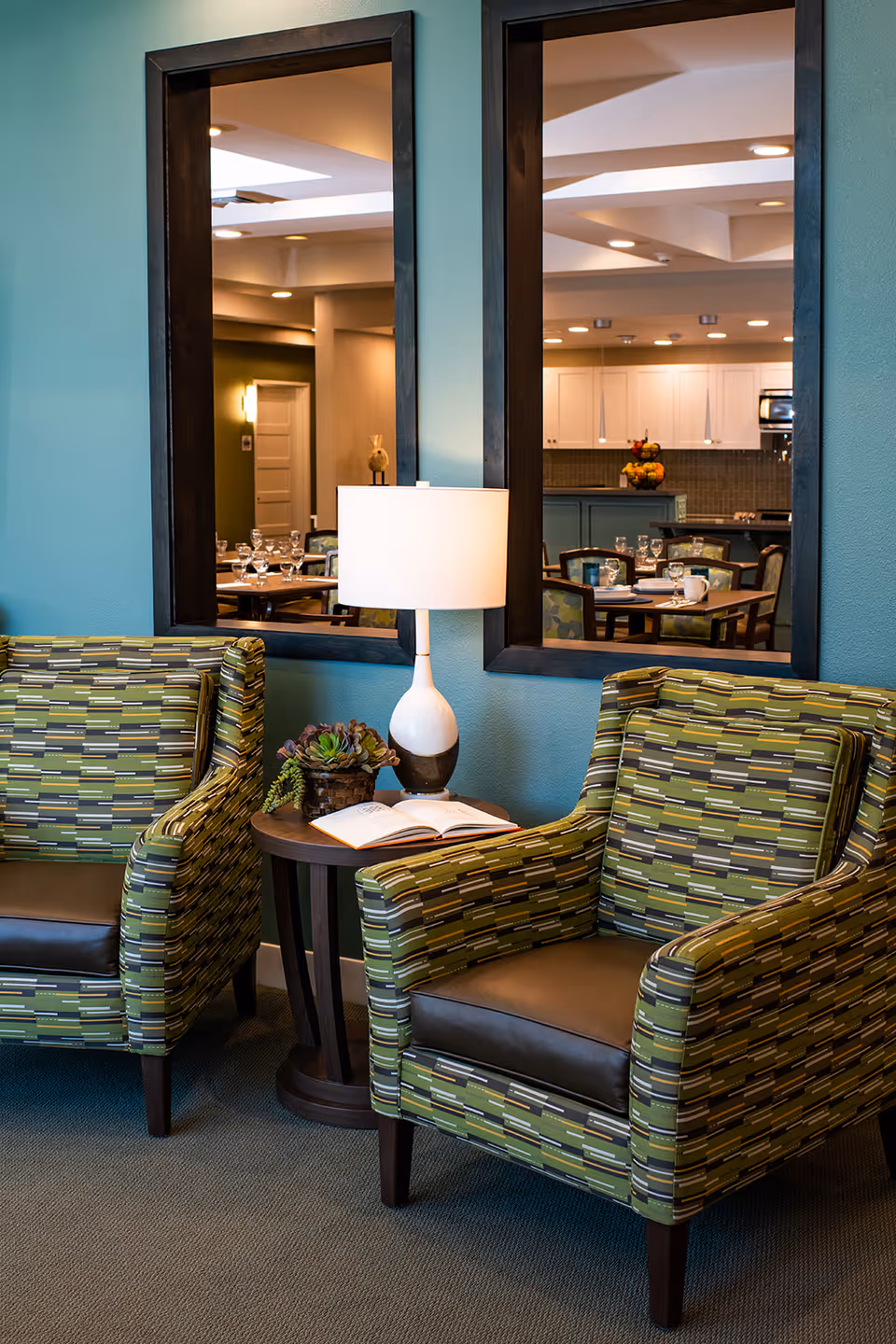 Two patterned armchairs with green, brown, and beige stripes are placed on either side of a small round wooden table. On the table is a white lamp with a dark base, an open book, and a small succulent plant. Behind the chairs, two large rectangular mirrors hang on a teal-colored wall, reflecting a dining area with tables, chairs, and a kitchen in the background.