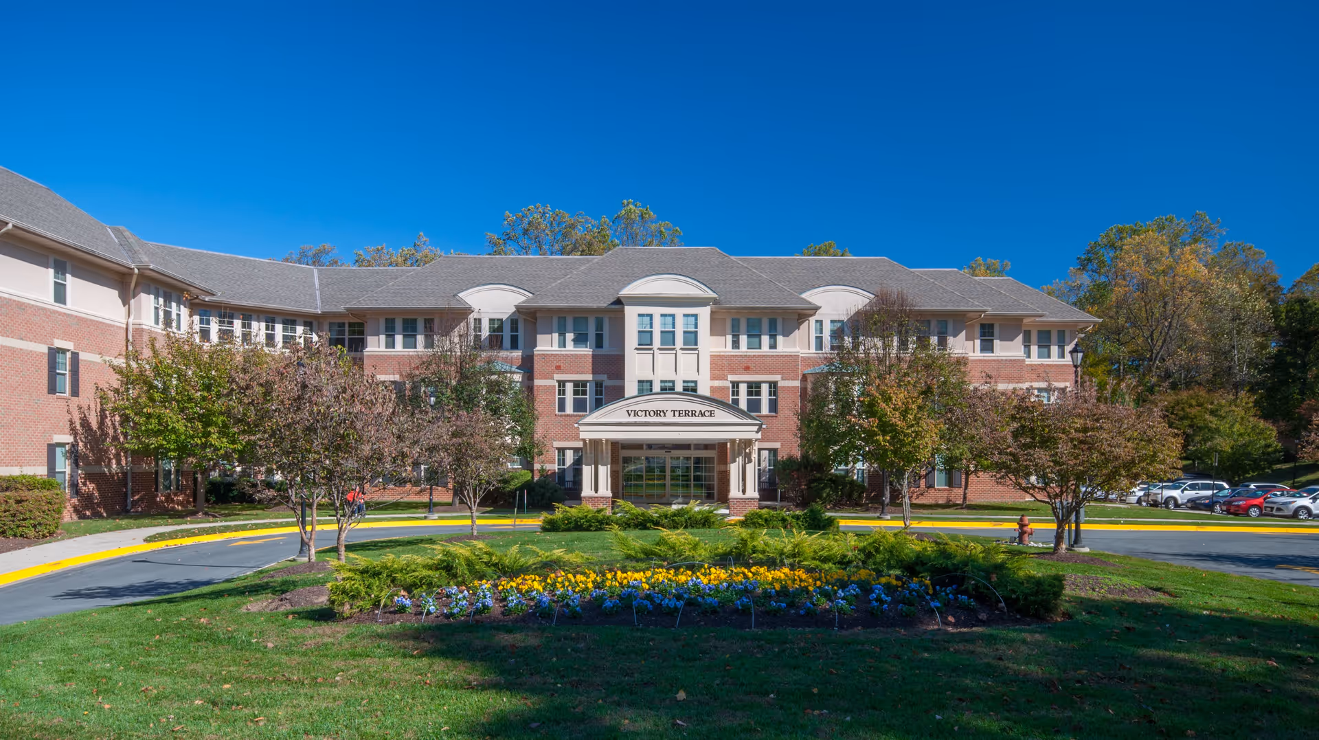 Front exterior view of Victory Terrace, a three-story senior living facility with a brick facade, multiple windows, and a covered entrance. The building is surrounded by trees, a circular driveway, and a landscaped flower bed with colorful flowers in the foreground under a clear blue sky.