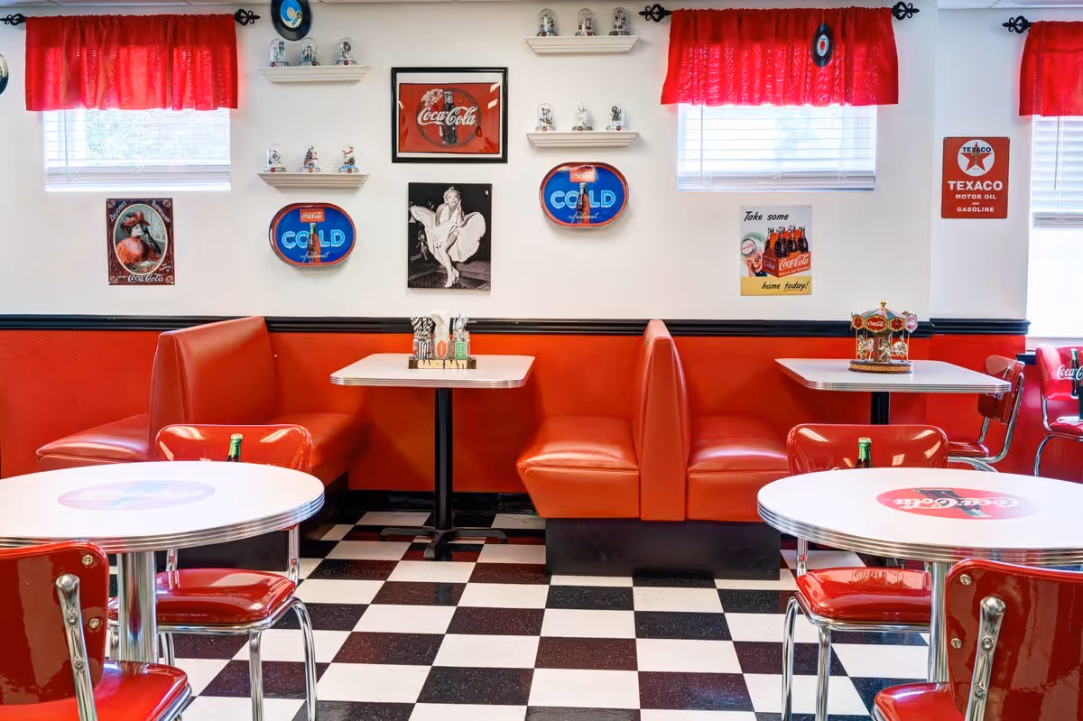 Retro-style dining area with red vinyl booths and chairs, white tables featuring Coca-Cola logos, black and white checkered floor, red curtains on windows, and vintage Coca-Cola and Texaco wall decorations.
