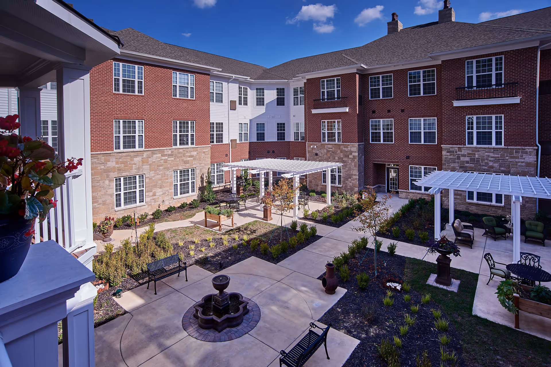 Outdoor courtyard area of a senior living facility with brick and stone exterior walls, multiple windows, paved walkways, benches, a small fountain, pergolas with seating areas, and landscaped plants and trees under a clear blue sky.