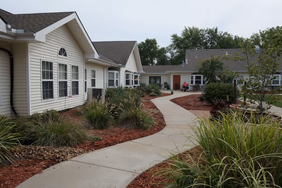 A paved walkway curves through a landscaped garden area with bushes and ornamental grasses, leading to a single-story building with beige siding and multiple windows. There are trees and outdoor seating visible near the building under an overcast sky.