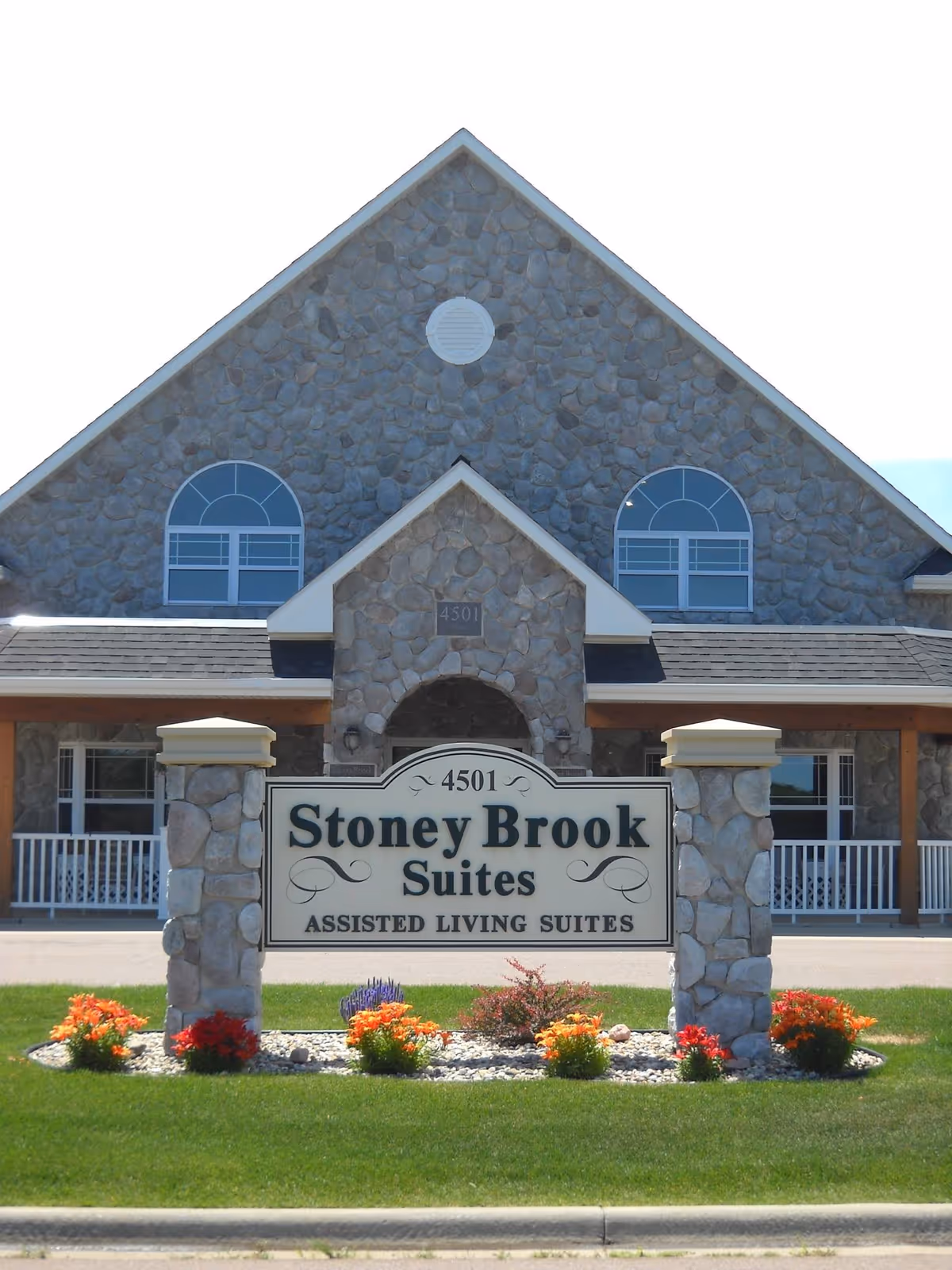 Front exterior view of Stoney Brook Suites Assisted Living building with a stone facade, two arched windows, and a sign in front surrounded by colorful flowers and green grass.