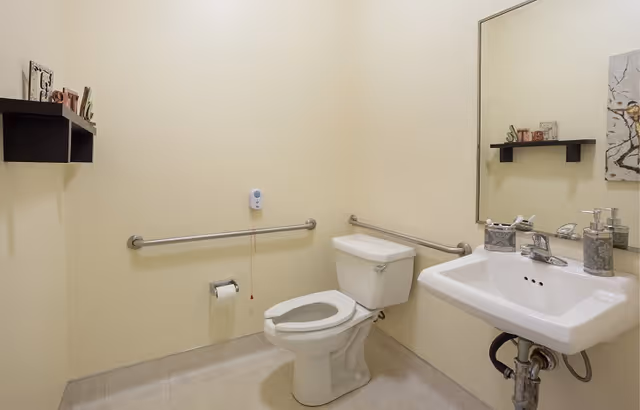 A clean, accessible bathroom with beige walls and tiled floor. The room features a white toilet with grab bars on both sides, a white sink with a large mirror above it, and a small black wall shelf holding decorative items. There is a wall-mounted emergency pull cord near the toilet.