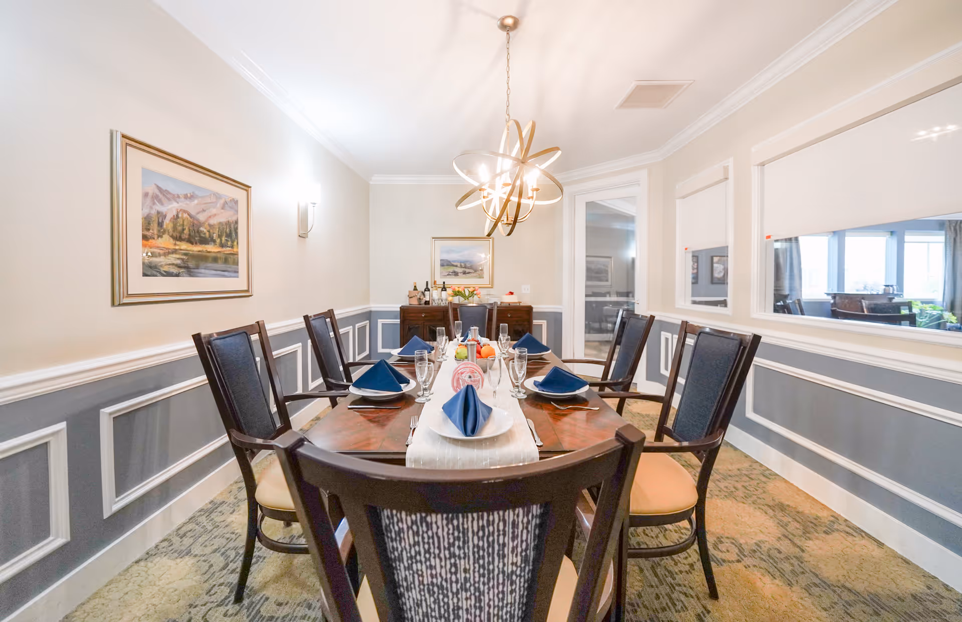 A well-lit dining room with a wooden dining table set for six people. The table has blue folded napkins, plates, glasses, and silverware. The room features gray and white wainscoting on the walls, a modern chandelier hanging above the table, framed landscape paintings on the walls, and a sideboard with bottles and flowers in the background. Large windows on the right side show another room with seating.