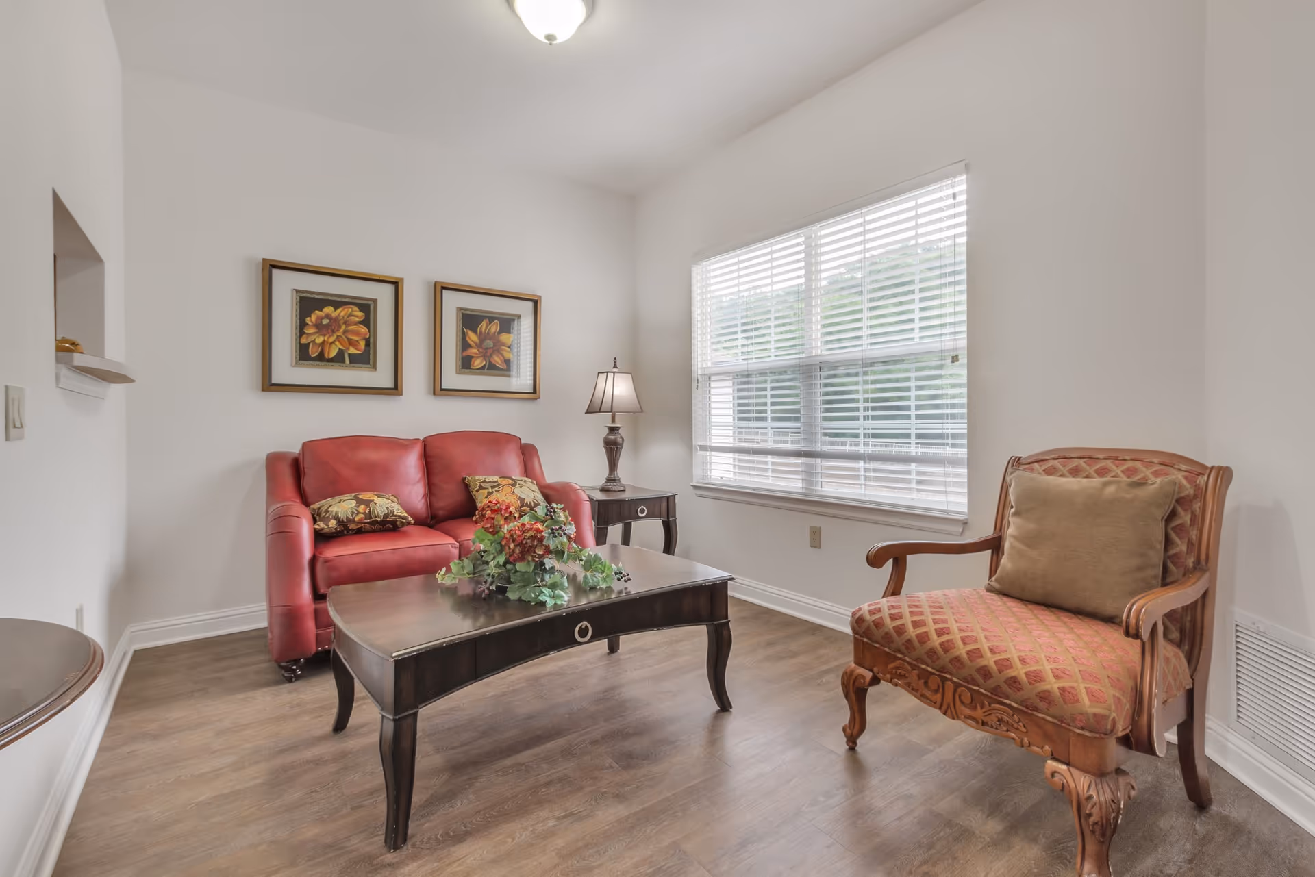 Small living room with a red loveseat, patterned armchair, coffee table with a floral arrangement, a lamp, and a large window with blinds.