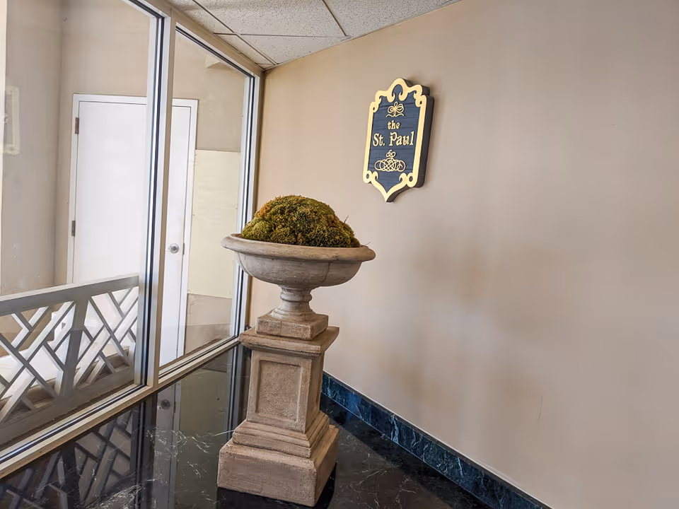 An indoor corner area with a decorative stone pedestal holding a moss-covered arrangement. A glass window and door are on the left side, and a decorative sign on the beige wall reads 'the St. Paul' in gold lettering.
