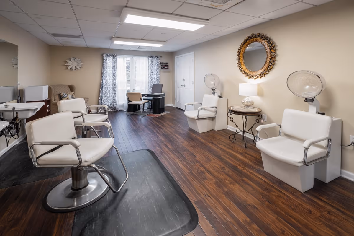 Interior view of a salon area with white salon chairs, hair dryers, a large ornate round mirror on the wall, a small round table with a lamp, and a desk with chairs near a window with patterned curtains. The floor is wood and the walls are beige.