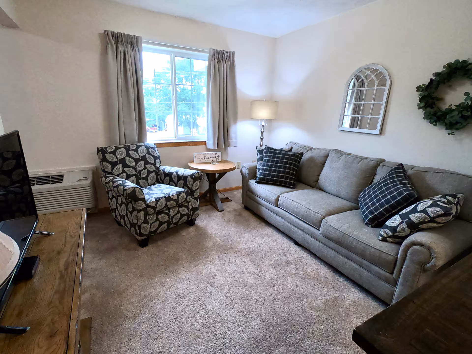 A cozy living room with a gray sofa adorned with patterned pillows, a matching armchair with leaf designs, a round wooden side table with a decorative 'family' sign, a floor lamp, a window with beige curtains, a wall mirror, and a green wreath on the wall.
