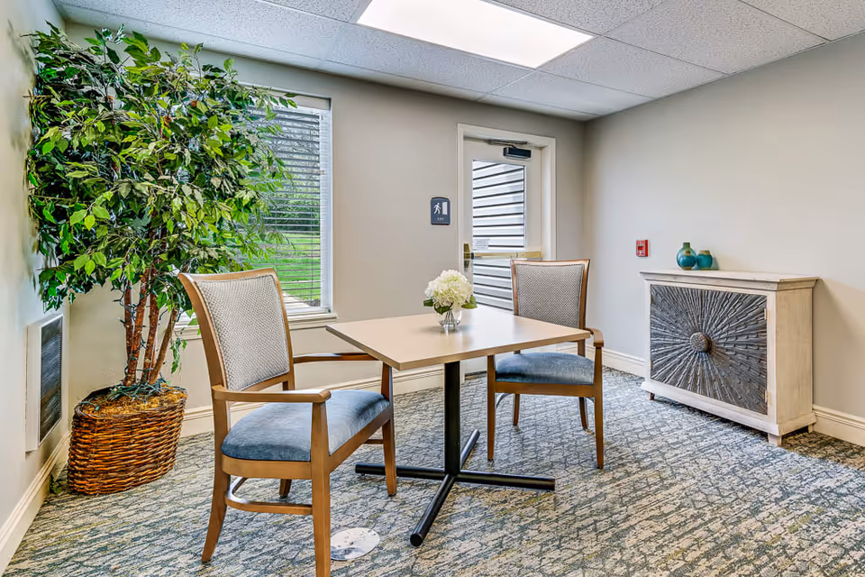 A small seating area in a senior living facility with a square table and two wooden chairs with blue cushions. There is a potted artificial tree in a wicker basket near a window with blinds. A decorative cabinet with two vases on top is against the wall. A door with a sign indicating an exit is visible in the background.
