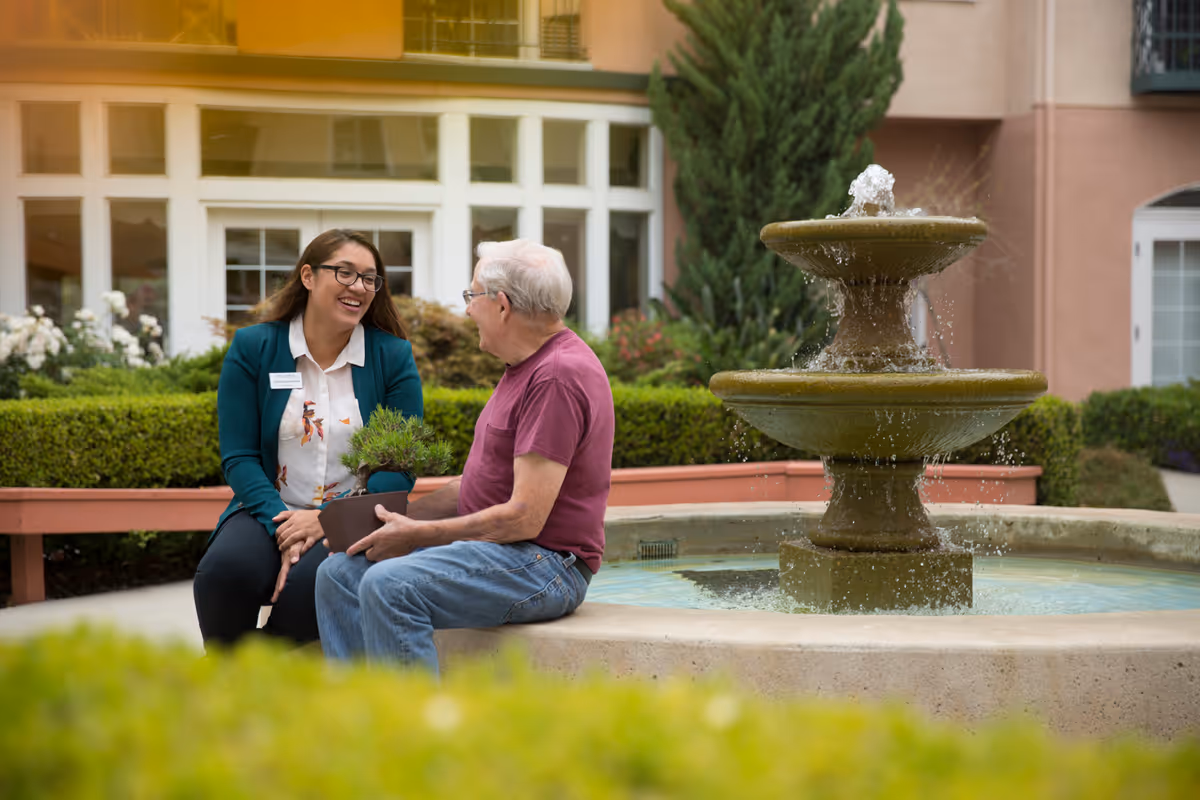 An elderly man and a woman with a name tag sit and chat happily beside a stone water fountain in a garden courtyard with greenery and flowers around them.