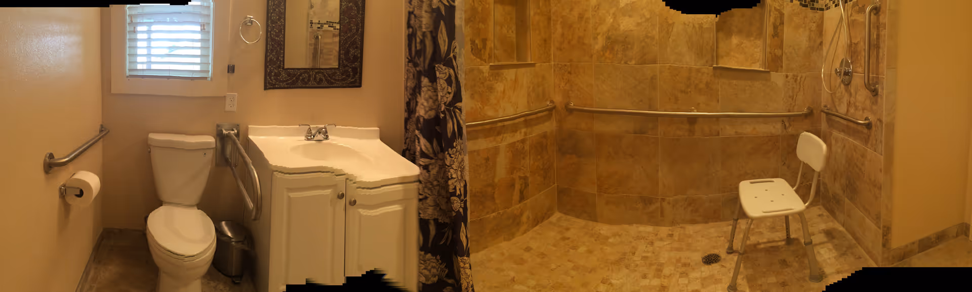 A bathroom with beige walls and tiled floors featuring a white toilet with grab bars on both sides, a white sink with a cabinet underneath and a mirror above, and a walk-in shower area with beige tiles, grab bars, and a white shower chair.