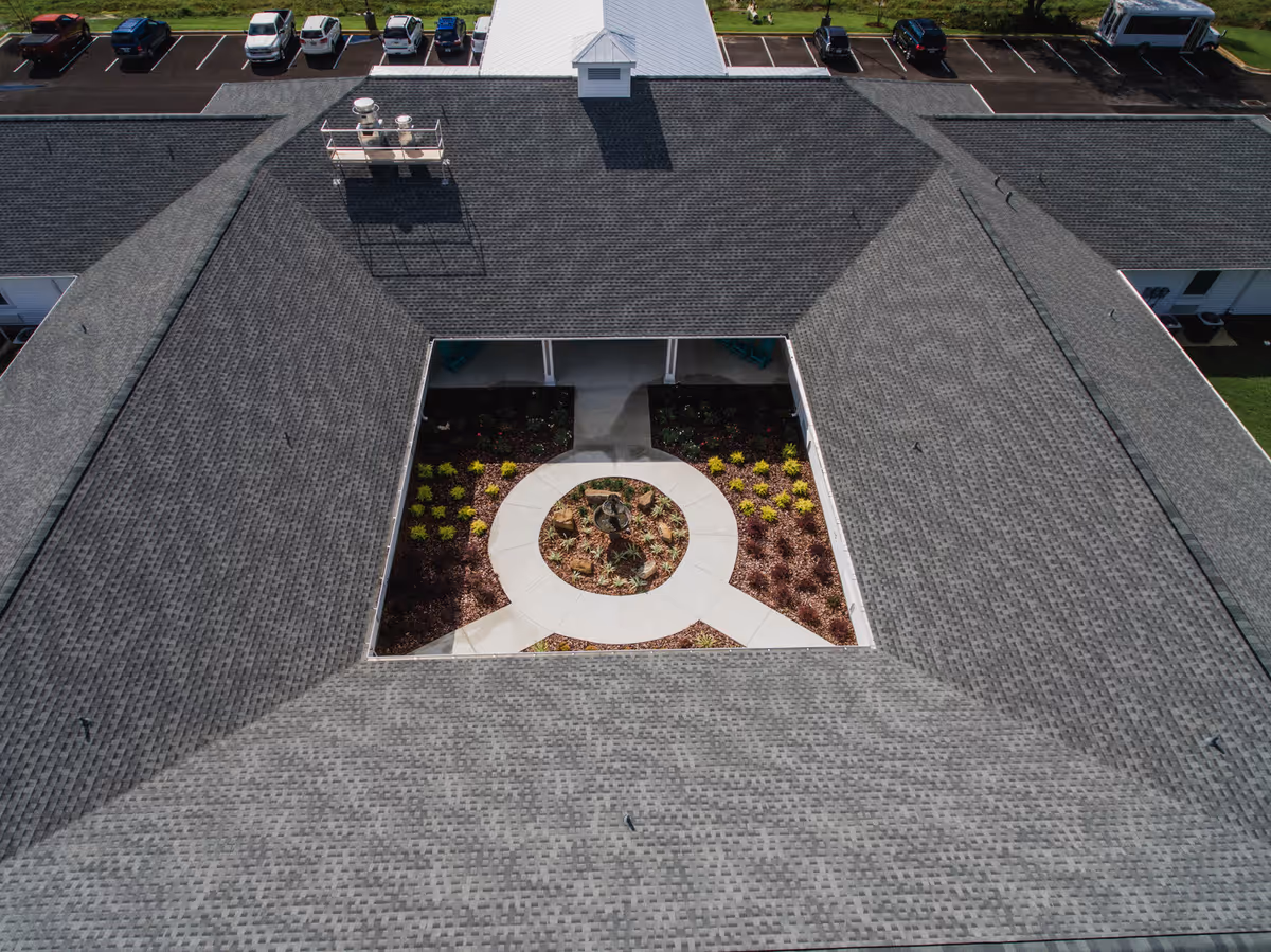 Aerial view of a senior living facility named Navarre Gardens showing a large building with a central courtyard garden. The courtyard has a circular walkway surrounding a small garden with plants and a fountain in the center. The building has a gray shingled roof and parking spaces with several vehicles visible in the background.