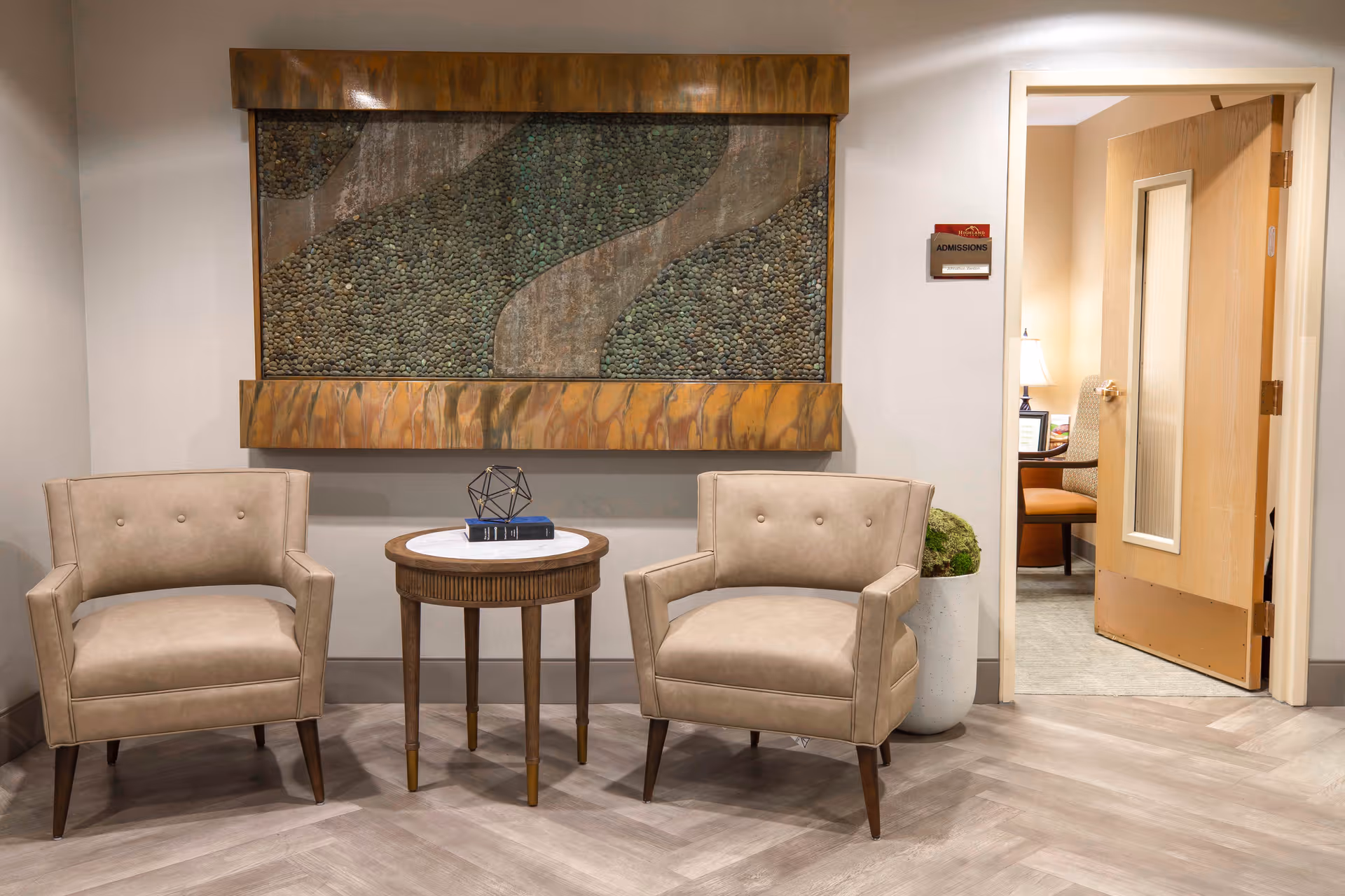 A waiting area with two beige armchairs and a small round wooden table between them. On the table are two books and a decorative geometric object. Behind the chairs is a large framed wall art piece featuring a textured design with stones and a flowing pattern. To the right, an open wooden door leads to another room with a lamp and a chair visible inside. A small sign labeled 'Admissions' is mounted on the wall next to the door.