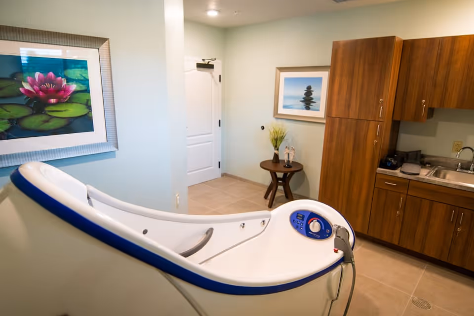 Interior bathing room with a white curved therapeutic tub in the foreground, wooden cabinets and sink on the right, and framed artwork on pale walls.