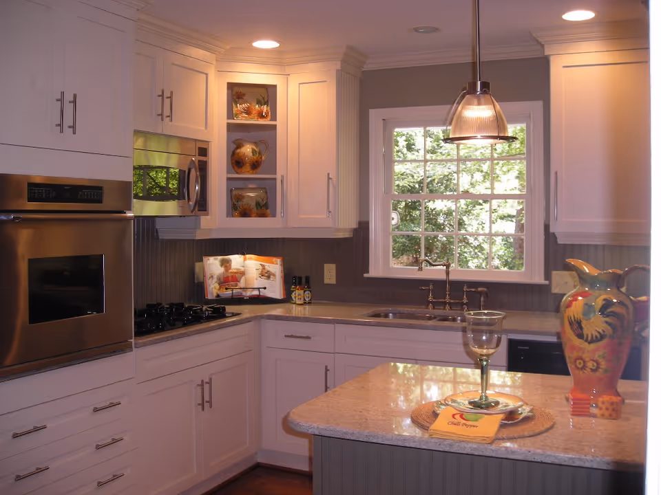 A modern kitchen with white cabinetry, stainless steel oven and microwave, a gas stove, and a window above the sink showing greenery outside. The countertop has a decorative pitcher, a wine glass on a placemat, and a cookbook on a stand.