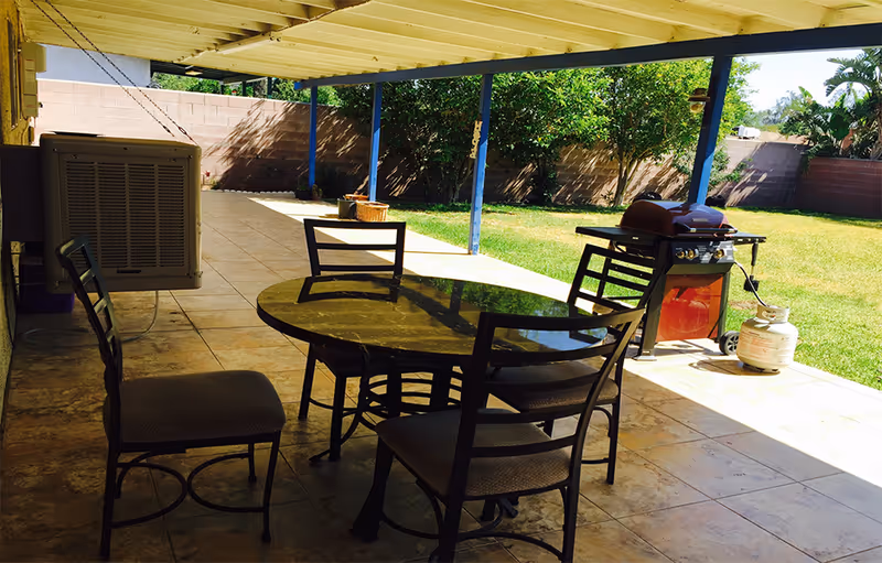 Covered outdoor patio area with a round glass table and four metal chairs with cushions. There is a gas grill with a propane tank on the tiled floor. The patio overlooks a grassy backyard with trees and a brick wall fence.