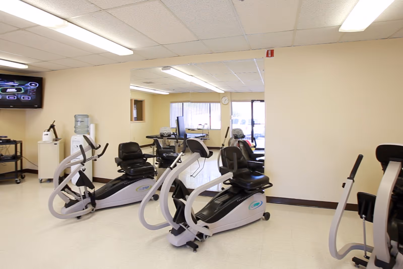 Interior view of a fitness room with several recumbent exercise bikes arranged on a light-colored floor. The room has beige walls, fluorescent ceiling lights, a wall-mounted TV, a water cooler, and a clock on the far wall near windows with vertical blinds.