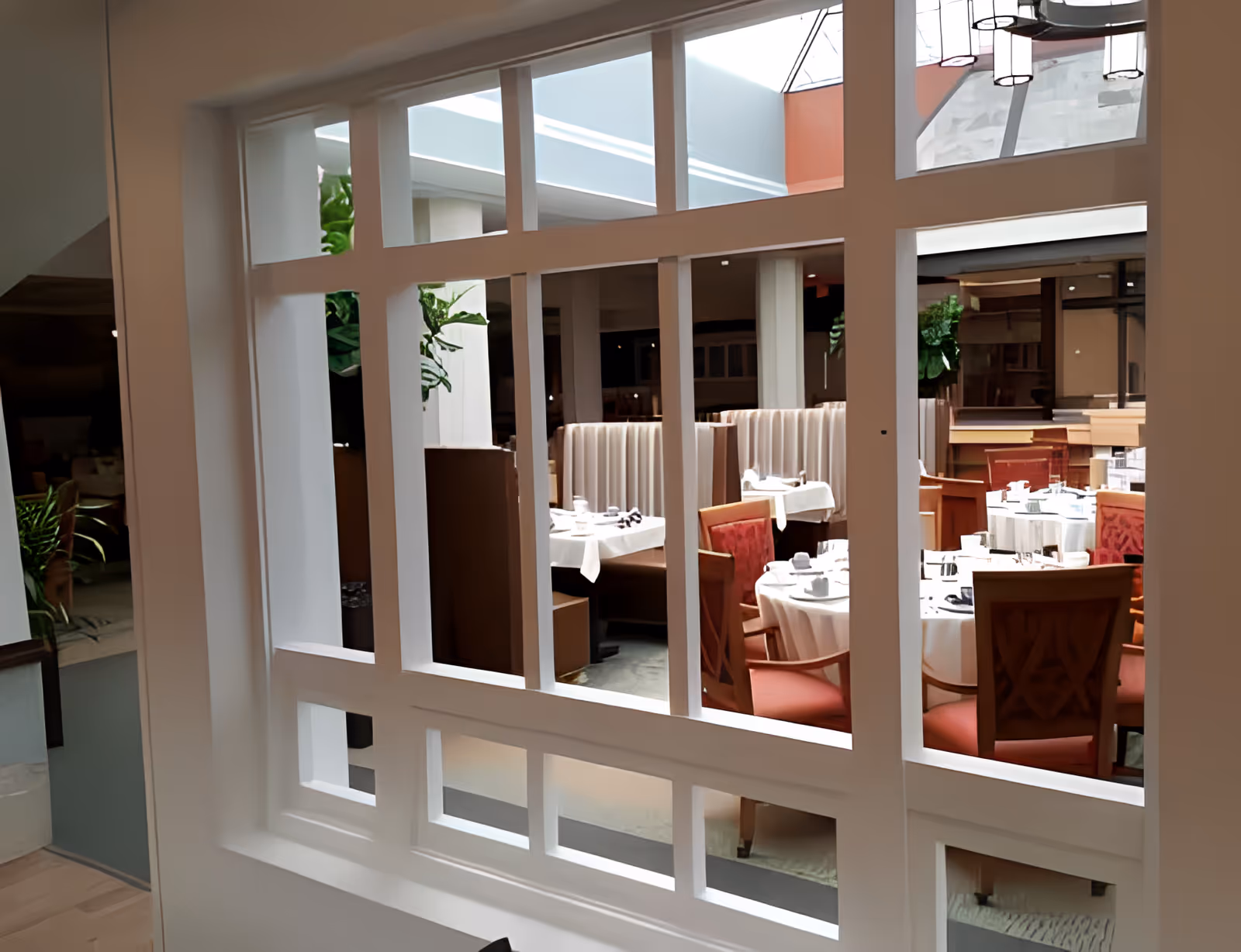 View through a white decorative partition into a dining room with round tables set for service, red chairs and booth seating.