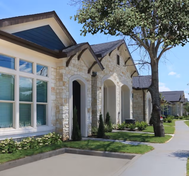 Exterior view of a luxury active-adult community building with stone facade, large windows, arched doorways, and a tree-lined sidewalk under a clear blue sky.