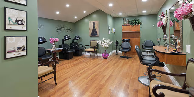 Interior view of a salon area with green walls and wooden flooring. The room features multiple black salon chairs, hair drying stations, and mirrors with floral decorations. There are framed pictures on the walls and vases with flowers placed around the room.