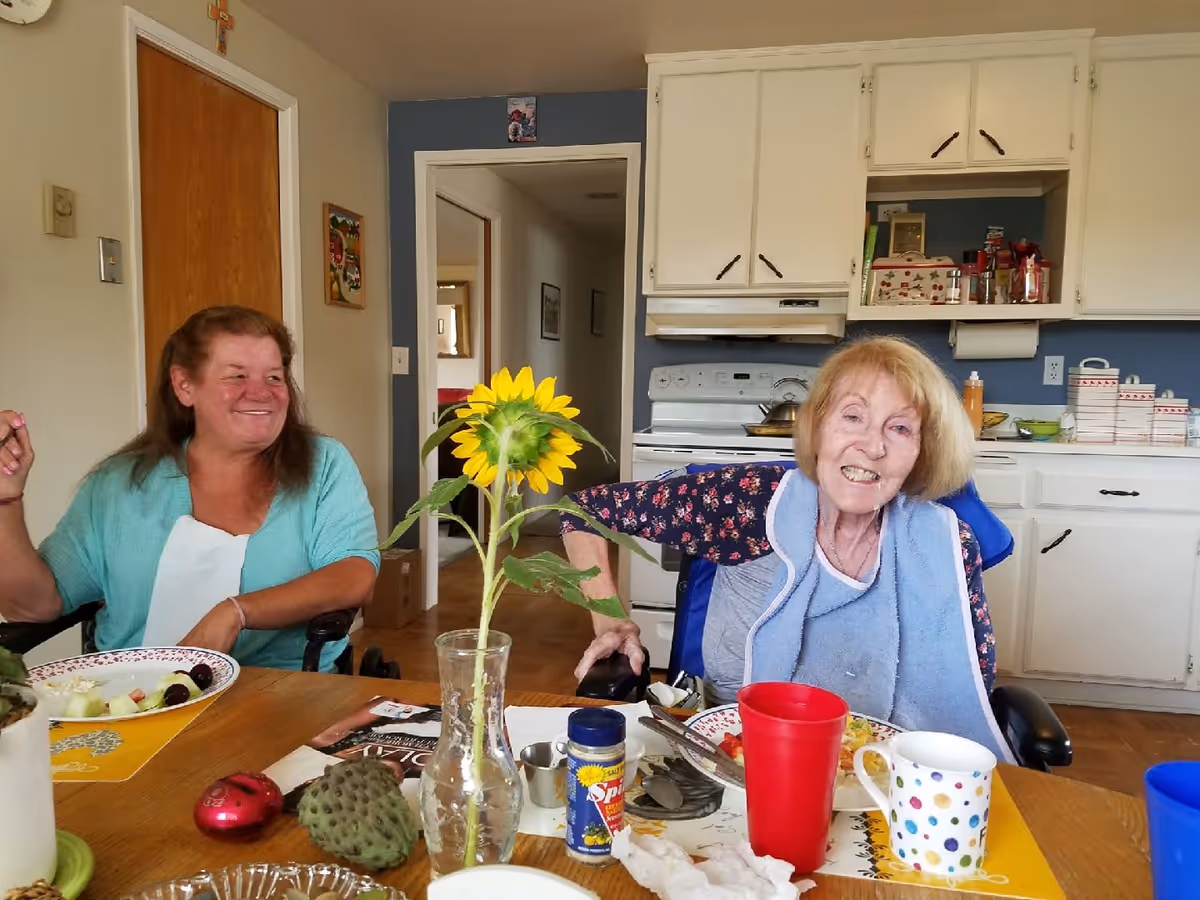 Two women sitting at a dining table in a kitchen. One woman is wearing a light blue top and smiling, while the other woman, wearing a floral top and a blue bib, is also smiling. On the table are plates with food, a vase with a sunflower, cups, and various condiments. The background shows white kitchen cabinets, a stove, and some kitchen items.