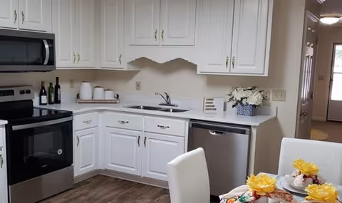 A modern kitchen with white cabinets, a stainless steel oven, microwave, and dishwasher. The countertop has a sink, some decorative jars, and a vase with white flowers. In the foreground, there is a dining table set with white chairs and yellow flower decorations on the plates.