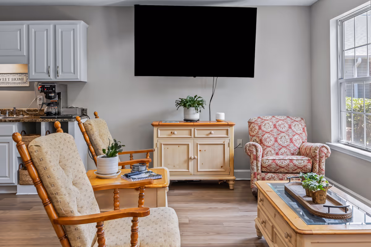 A cozy living room area with two cushioned wooden armchairs, a patterned upholstered armchair, a wooden coffee table with a plant centerpiece, a wooden cabinet with a potted plant and a small speaker on top, a large flat-screen TV mounted on the wall, and a window letting in natural light. A small kitchen area with white cabinets and a coffee maker is visible in the background.