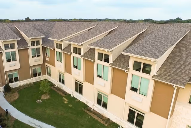 Exterior view of a multi-story senior living facility building with beige and brown walls, multiple windows, and a shingled roof. There is a green lawn and a paved walkway in front of the building.