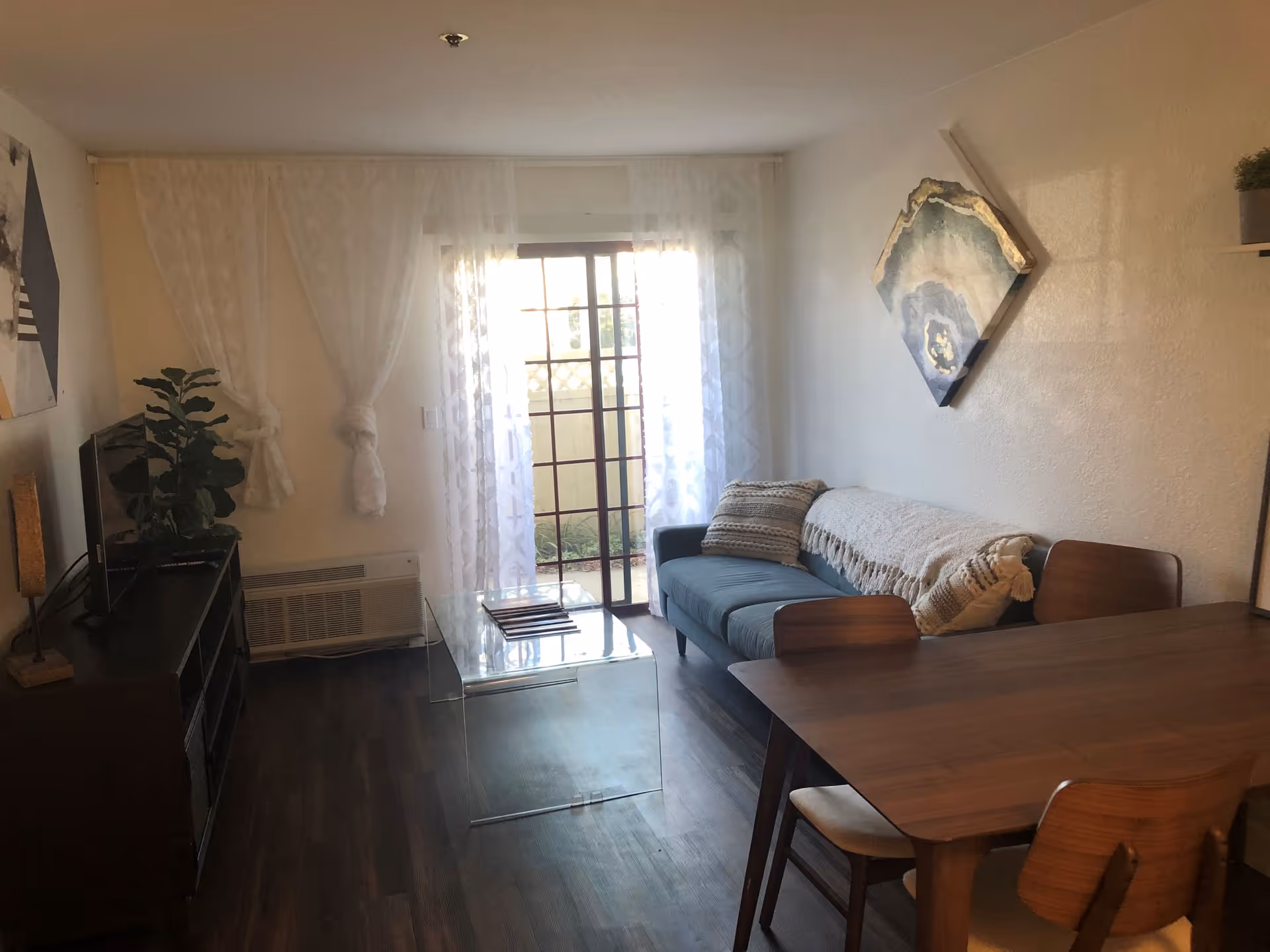 A cozy living room with a dark wooden floor, a blue sofa with cushions and a throw blanket, a clear glass coffee table, a wooden dining table with four chairs, a TV on a dark wooden stand, a potted plant, and sheer white curtains covering a sliding glass door leading outside.