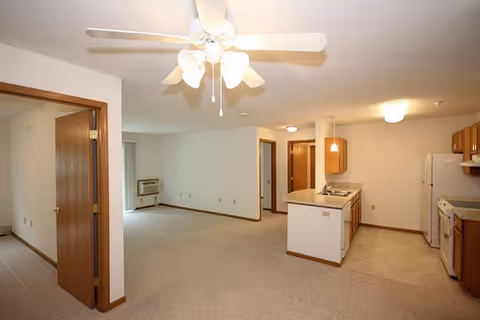 Interior view of an unfurnished senior apartment showing an open living area with beige carpet, a ceiling fan with lights, a kitchen with wooden cabinets, white appliances including a refrigerator and stove, and an open doorway leading to another room.
