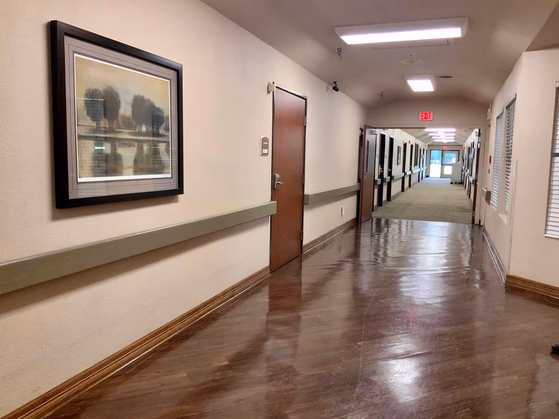 Long polished interior hallway in a care facility with room doors, handrails, framed artwork, and an exit sign at the far end.