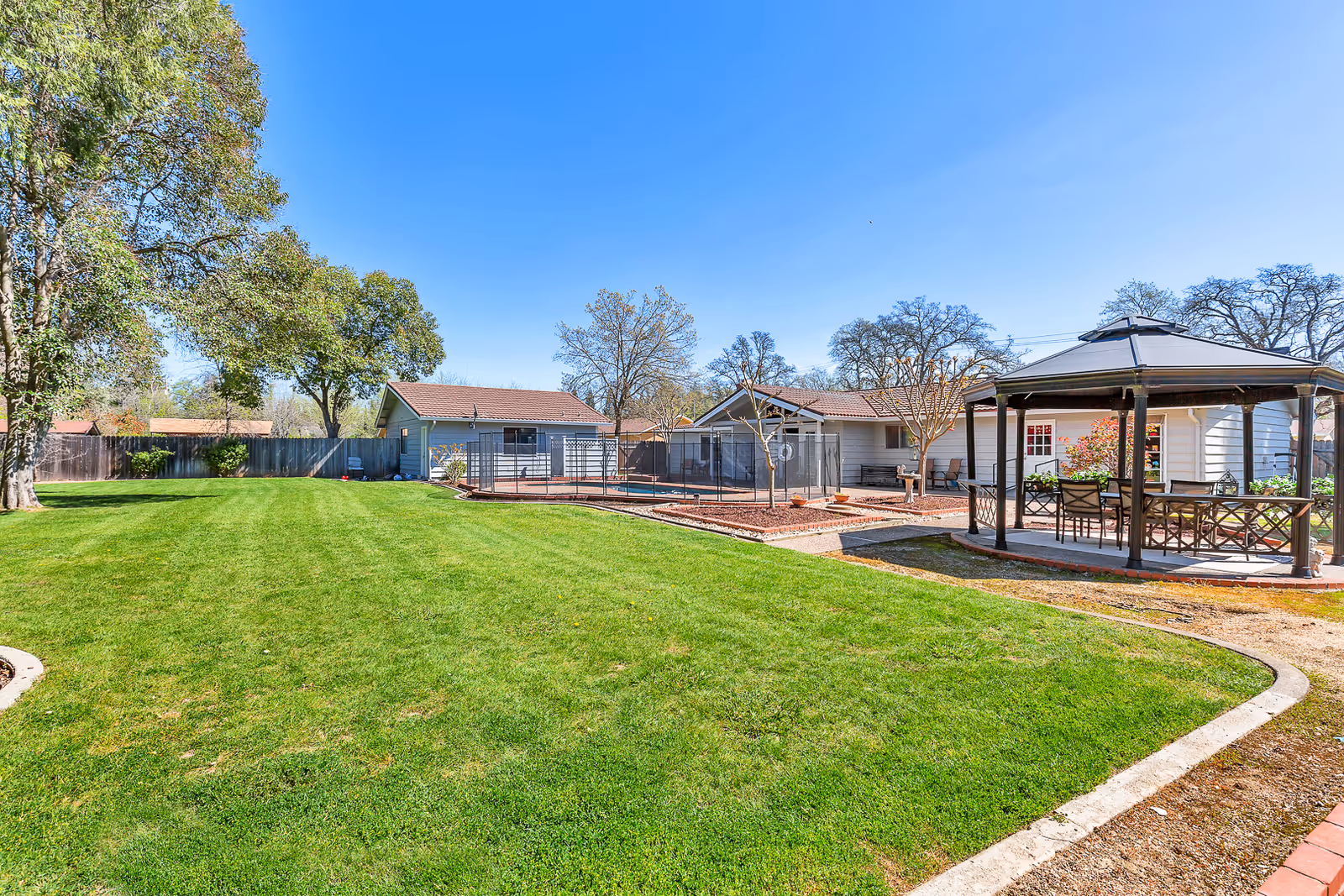 A spacious outdoor area with a well-maintained green lawn, several trees, and a gazebo with chairs and a table. In the background, there are single-story buildings with red-tiled roofs and a fenced area. The sky is clear and blue.