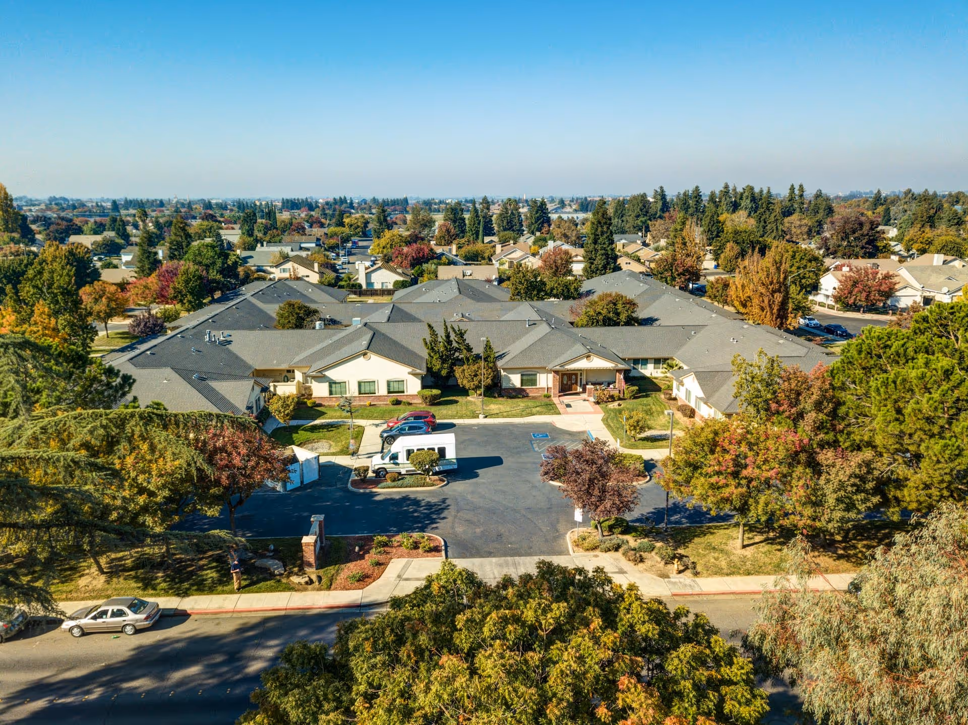 Aerial view of a senior living facility named Cogir of Turlock surrounded by trees and residential houses under a clear blue sky. The facility has multiple connected buildings with gray roofs and a parking area with a few vehicles.