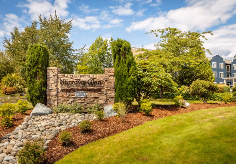 Stone sign for Narrows Glen surrounded by landscaped greenery including bushes, trees, and rocks, with a multi-story building partially visible in the background under a partly cloudy sky.