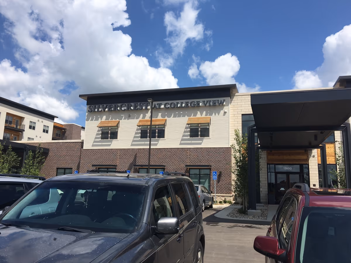 Exterior front entrance of the Silvercrest at College View senior living building with parked cars and a canopy under a partly cloudy blue sky.