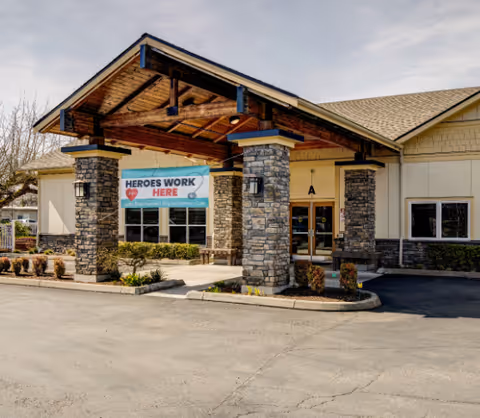 Entrance of Spring Ridge Assisted Living and Memory Care facility with a covered driveway supported by stone pillars. A banner reading 'HEROES WORK HERE' is displayed above the entrance. The building has beige siding and a shingled roof, with some landscaping around the entrance.