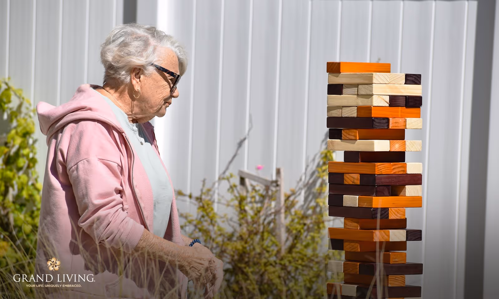 An elderly woman wearing glasses and a pink hoodie is playing a large outdoor Jenga game with wooden blocks in various shades of brown, orange, and beige. She is standing outside near some plants and a white fence.