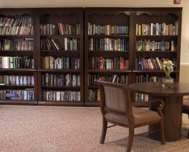 Wooden bookshelves filled with books line the wall beside a round table and chairs in a carpeted reading area.