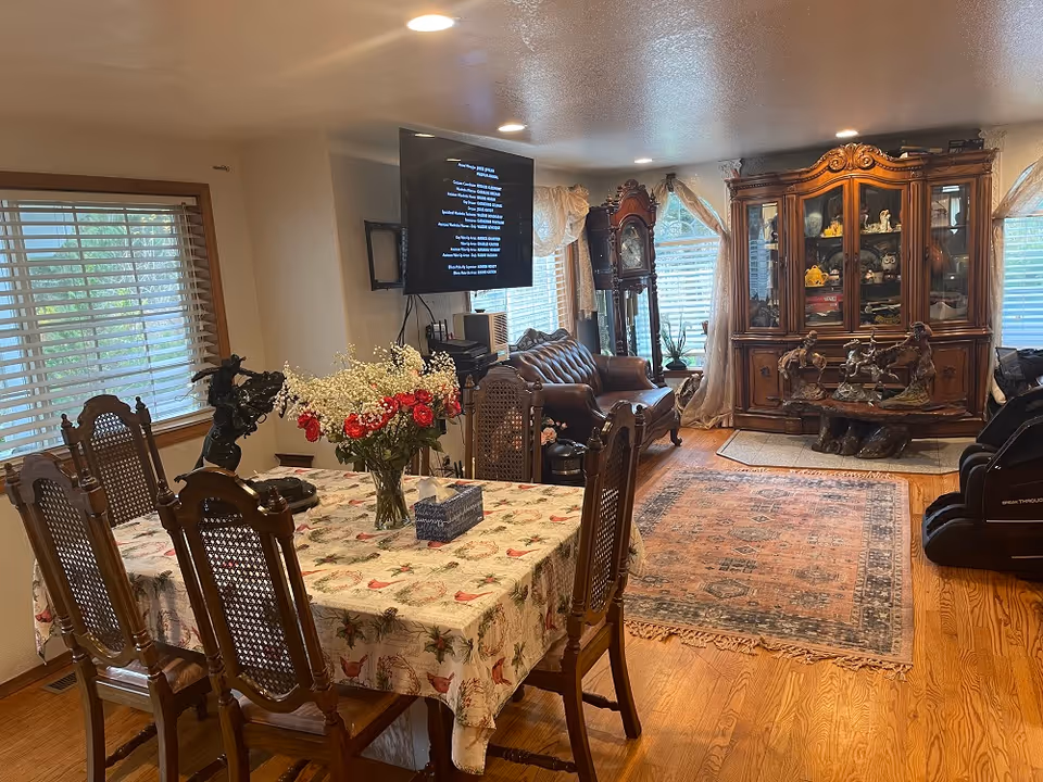 A cozy living and dining area in Tumwater Meadows Adult Family Home featuring a wooden dining table with six chairs and a floral tablecloth, a vase with red and white flowers, a mounted flat-screen TV, a brown leather sofa, a grandfather clock, a large wooden display cabinet with glass doors, decorative horse sculptures, and large windows with blinds and curtains letting in natural light.