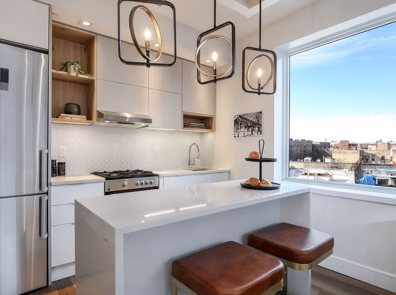 Modern kitchen with white cabinetry, stainless steel refrigerator and stove, a white island with two brown cushioned stools, three pendant lights hanging above, and a large window showing an urban cityscape outside.