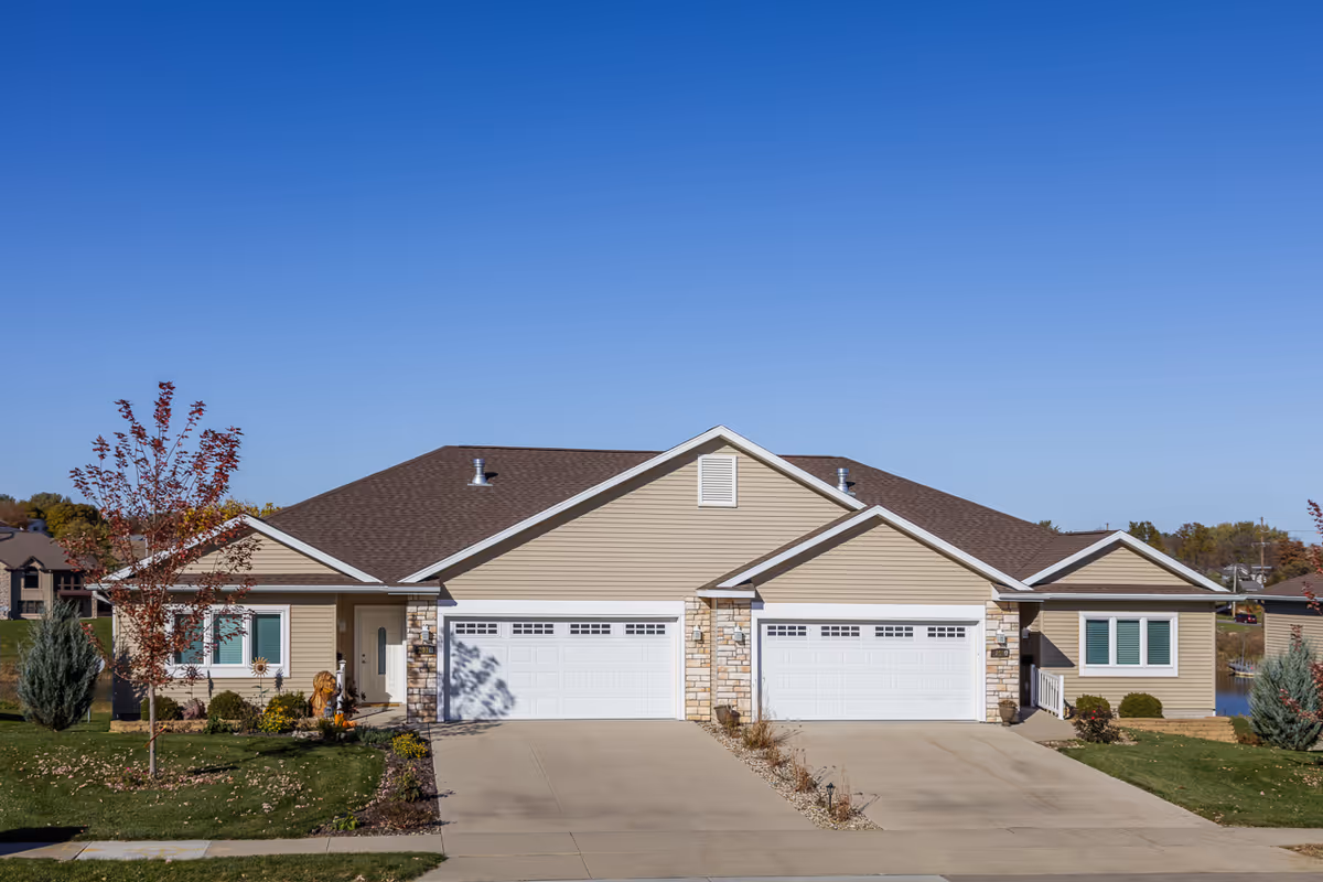 Exterior view of a beige duplex building with two white garage doors, a brown shingled roof, and small landscaped front yards with trees and shrubs under a clear blue sky.
