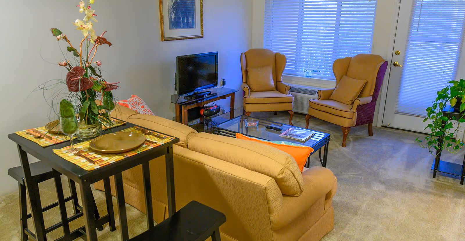 A cozy living room with a beige sofa, two beige armchairs with pillows, a glass coffee table with a remote and magazine, a small TV on a wooden stand, a tall plant on a black side table, and a small dining table set for two with placemats, plates, and a decorative flower arrangement. The room has carpeted flooring, a window with blinds, and a door with a window also covered by blinds.
