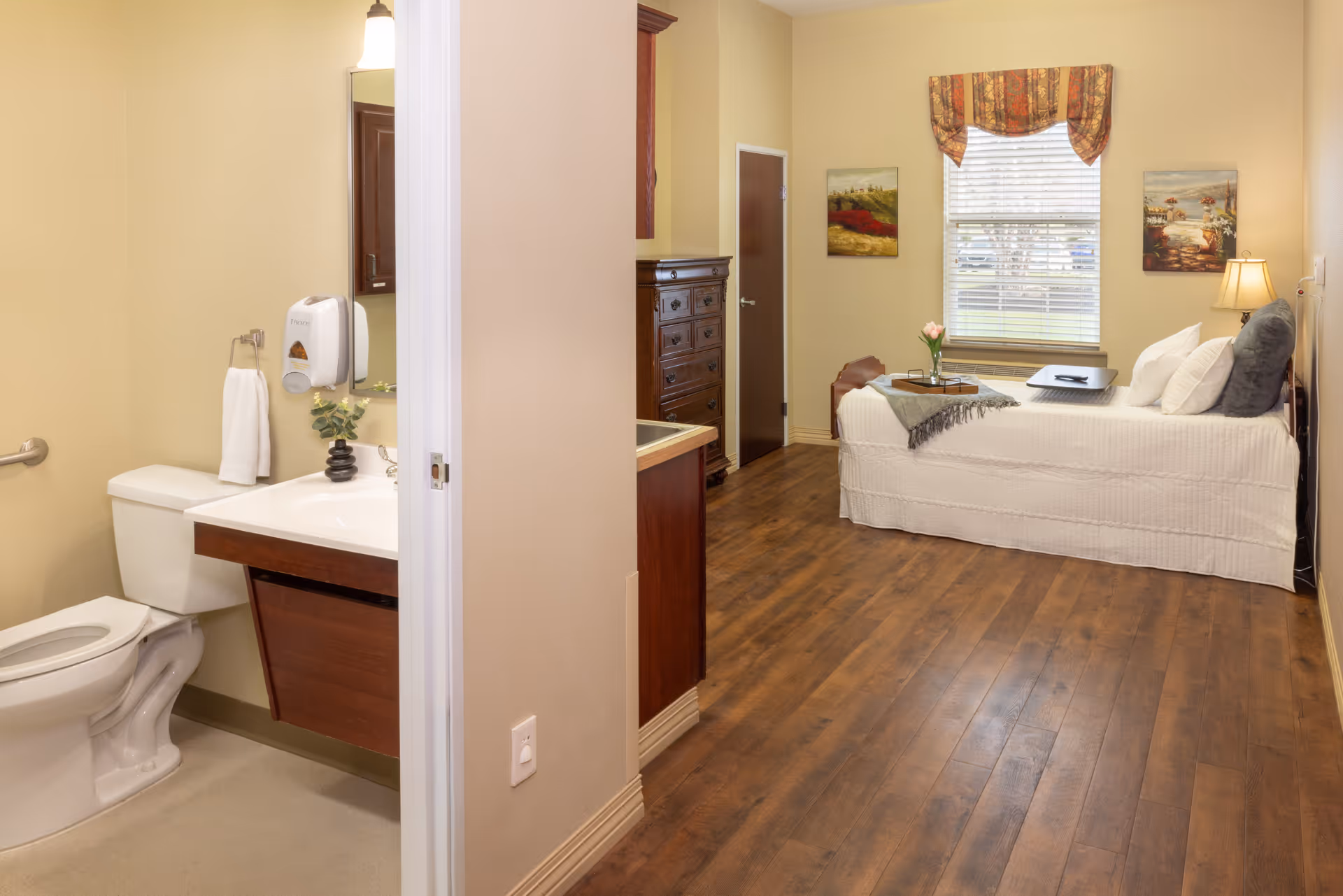 A furnished senior living bedroom with a bed, dresser, window and an adjoining accessible bathroom visible to the left.