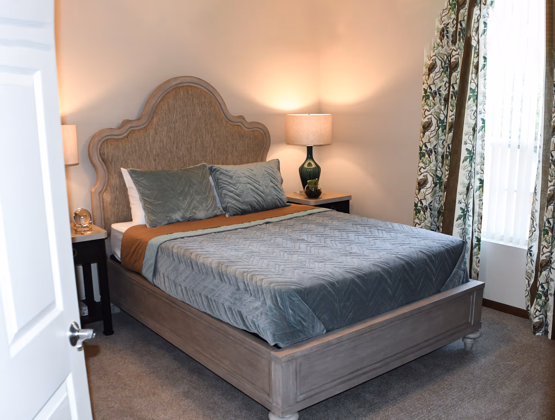 A cozy bedroom with a wooden bed frame and an upholstered headboard. The bed is made with a gray quilted bedspread, two gray pillows, and a brown blanket. There are two bedside tables, each with a lamp, and a window with floral patterned curtains letting in natural light.