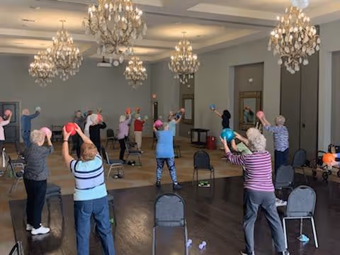 A group of elderly individuals participating in a seated exercise class in a spacious room with wooden floors and multiple chandeliers. They are holding colorful exercise balls above their heads while standing behind chairs.