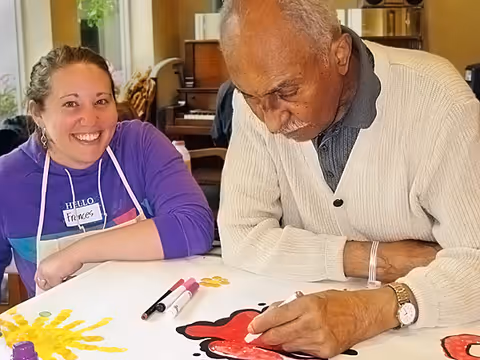 An elderly man coloring a red flower on a large sheet of paper with markers while a smiling woman wearing a purple shirt and a name tag that says 'Frances' watches him. They are seated at a table in a well-lit room with a piano and other furniture in the background.